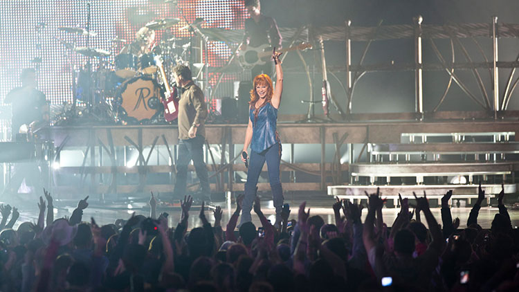 Country music singer Reba McEntire on stage singing a song with the crowd in front of her with her hand up, dancing.